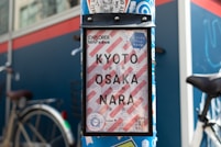 A signage indicating directions to Kyoto, Osaka, and Nara is mounted on a street pole. The sign has a striped red and white background with bold black letters. A bicycle is partially visible in the background, alongside a blue wall adorned with various stickers and logos. The sign also mentions 'Explorer Map' and 'Guide Map' with additional text in both English and Japanese.
