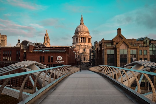 A wide-angle view of a modern pedestrian bridge leading towards an iconic cathedral with a large dome. Flanking the bridge are brick and glass buildings, one of which has the sign 'City of London School'. The sky is partly cloudy, casting a serene backdrop for the historic architecture.