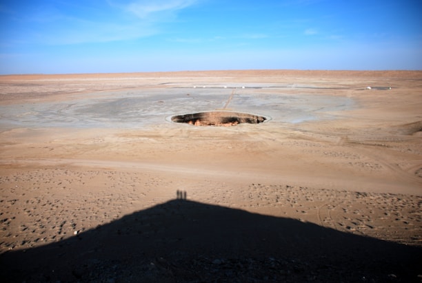 A vast desert landscape with a large circular hole in the ground. The area is mostly barren with patches of rough terrain and small scattered structures in the distance. The sky is clear and blue, casting strong sunlight that creates a shadow at the bottom of the image. Two small human silhouettes are visible in the shadow, suggesting the presence of observers.