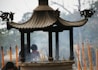 An ornate incense burner with a pagoda-style roof is surrounded by burning incense sticks, creating a smoky atmosphere. In the background, there are blurred figures and trees, suggesting a setting in a traditional or religious site.