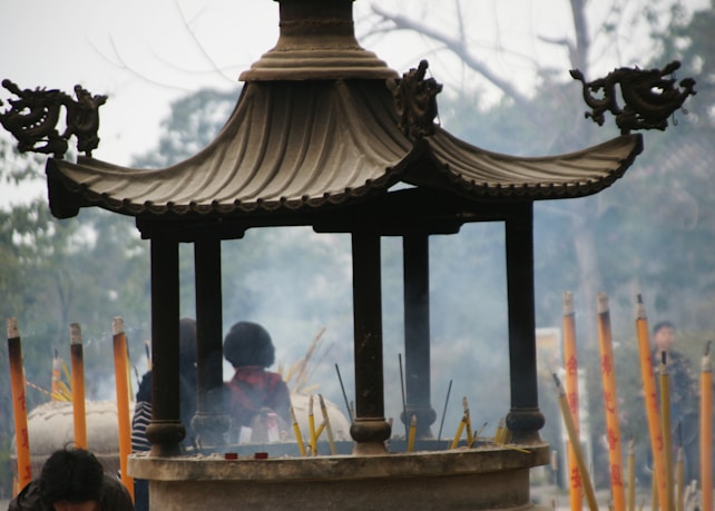 An ornate incense burner with a pagoda-style roof is surrounded by burning incense sticks, creating a smoky atmosphere. In the background, there are blurred figures and trees, suggesting a setting in a traditional or religious site.