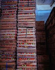 Stacks of wooden crates filled with colorful apples are arranged in a warehouse. The crates are piled high, each containing apples in shades of red, green, and yellow. The lighting is dim, casting shadows across the scene and highlighting the textures of the wooden crates.