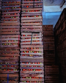 Stacks of wooden crates filled with colorful apples are arranged in a warehouse. The crates are piled high, each containing apples in shades of red, green, and yellow. The lighting is dim, casting shadows across the scene and highlighting the textures of the wooden crates.
