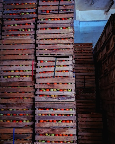 Stacks of wooden crates filled with colorful apples are arranged in a warehouse. The crates are piled high, each containing apples in shades of red, green, and yellow. The lighting is dim, casting shadows across the scene and highlighting the textures of the wooden crates.