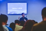 A person is speaking in front of an audience in a room with a screen displaying a biblical passage from Hebrews 11:32-40. The room appears to have subdued lighting with an emphasis on the speaker who is holding a microphone.