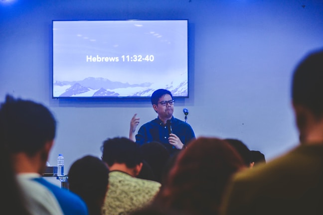 A person is speaking in front of an audience in a room with a screen displaying a biblical passage from Hebrews 11:32-40. The room appears to have subdued lighting with an emphasis on the speaker who is holding a microphone.