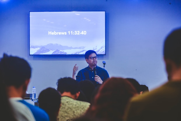 A person is speaking in front of an audience in a room with a screen displaying a biblical passage from Hebrews 11:32-40. The room appears to have subdued lighting with an emphasis on the speaker who is holding a microphone.