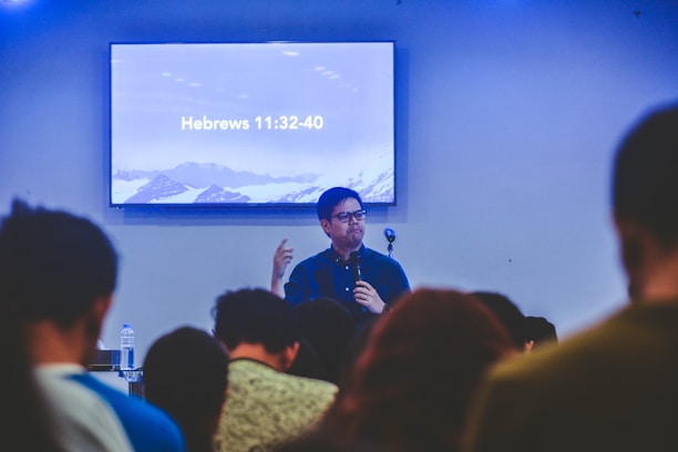A person is speaking in front of an audience in a room with a screen displaying a biblical passage from Hebrews 11:32-40. The room appears to have subdued lighting with an emphasis on the speaker who is holding a microphone.