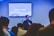 A person is speaking in front of an audience in a room with a screen displaying a biblical passage from Hebrews 11:32-40. The room appears to have subdued lighting with an emphasis on the speaker who is holding a microphone.