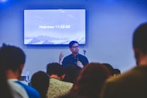 A person is speaking in front of an audience in a room with a screen displaying a biblical passage from Hebrews 11:32-40. The room appears to have subdued lighting with an emphasis on the speaker who is holding a microphone.