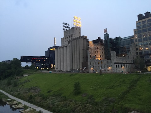 An industrial building complex with a historic stone structure featuring multiple silos and a large sign with bold yellow lettering on top. Surrounding the complex is a green grassy area with trees, and a concrete path runs alongside a river. The sky is overcast, lending a somewhat somber and muted atmosphere to the scene.