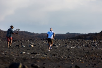A person jogs across a rocky, barren landscape with another person in pursuit holding a camera stabilizer. The terrain is rough and covered with large stones, under an open sky. The individuals are dressed for outdoor activity and appear to be engaged in capturing footage.