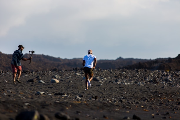 A person jogs across a rocky, barren landscape with another person in pursuit holding a camera stabilizer. The terrain is rough and covered with large stones, under an open sky. The individuals are dressed for outdoor activity and appear to be engaged in capturing footage.