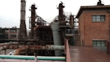 An industrial area with large metal structures and machinery surrounding a brick building with small windows. Rusted pipes and staircases are visible, indicating an old or unused factory setting.