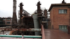 An industrial area with large metal structures and machinery surrounding a brick building with small windows. Rusted pipes and staircases are visible, indicating an old or unused factory setting.