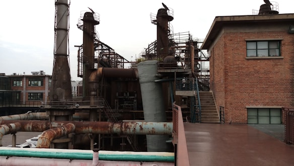 An industrial area with large metal structures and machinery surrounding a brick building with small windows. Rusted pipes and staircases are visible, indicating an old or unused factory setting.
