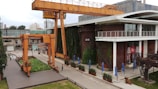 An industrial setting with large orange cranes positioned near a modern building. The building, labeled A18, has a vertical garden wall and features large windows alongside outdoor seating with blue umbrellas. The surrounding area includes industrial equipment and greenery.
