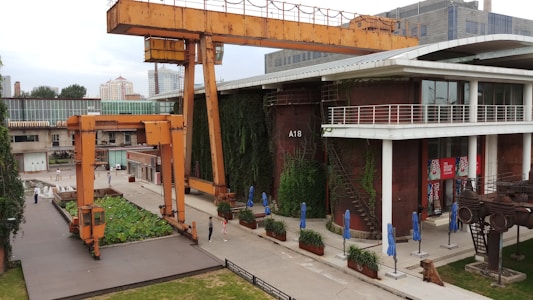 An industrial setting with large orange cranes positioned near a modern building. The building, labeled A18, has a vertical garden wall and features large windows alongside outdoor seating with blue umbrellas. The surrounding area includes industrial equipment and greenery.