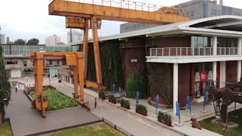 An industrial setting with large orange cranes positioned near a modern building. The building, labeled A18, has a vertical garden wall and features large windows alongside outdoor seating with blue umbrellas. The surrounding area includes industrial equipment and greenery.