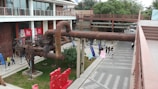 An industrial-style outdoor space featuring large rusted pipes and a design store with signage. Several people walk along the concrete path next to greenery and a bold red art installation. Overhead walkways and a modern building with glass windows are visible in the background.
