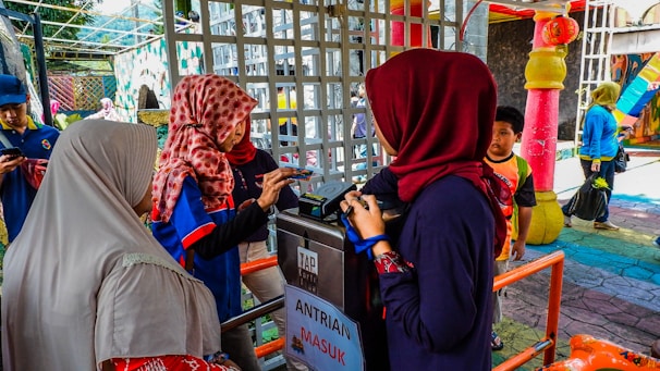 Several people are standing near a ticket or entry gate, engaging with an access control terminal. They are wearing colorful clothing, including hijabs. The environment appears lively and colorful with bright decorations and structures in the background.