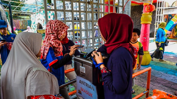 Several people are standing near a ticket or entry gate, engaging with an access control terminal. They are wearing colorful clothing, including hijabs. The environment appears lively and colorful with bright decorations and structures in the background.