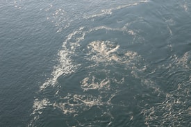 A close-up view of a large body of water with swirling currents and patterns created by waves. Sunlight reflects on the surface, creating shimmering highlights across the water.