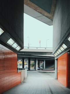 A modern urban underpass with concrete walls and ceiling. The pathway is bordered by orange tiled walls and illuminated by rectangular ceiling lights. Above the underpass, parts of a road bridge are visible with street signs and light poles.