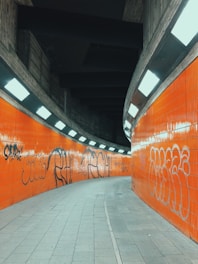 A curved underground pedestrian passageway with bright orange tiled walls, adorned with graffiti art. The ceiling is lined with fluorescent lights, creating a stark contrast against the dark concrete and enhancing the vibrant orange hue.
