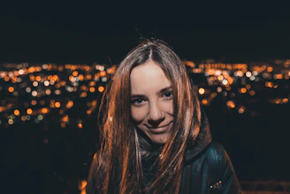 A warm photo of a smiling woman standing by a vibrant city nightscape with soft pastel lights.