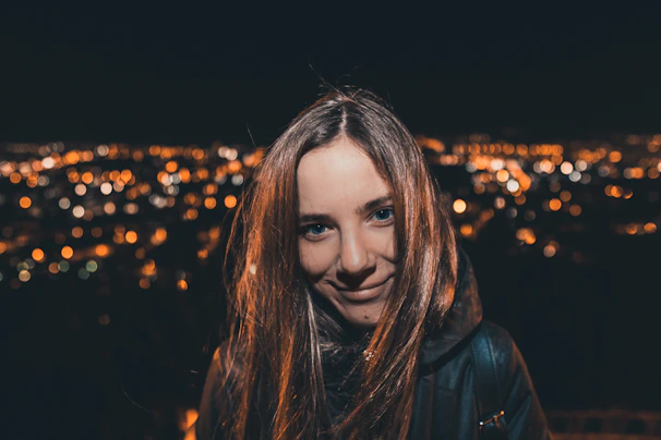 A warm photo of a smiling woman standing by a vibrant city nightscape with soft pastel lights.