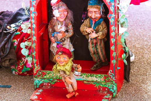 A colorful display featuring three puppets inside and around a red, ornately decorated booth. The puppets are dressed in vibrant clothing, with one female puppet wearing a floral dress and holding a mug, while another male puppet dons a striped outfit. A smaller, child-like puppet is positioned outside the booth, dressed in a polka-dot outfit with a red hat. The booth is decorated with green leafy designs.
