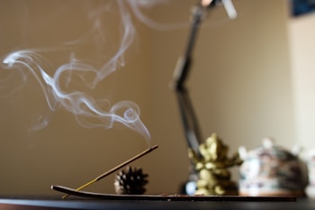 Wisps of smoke curl gracefully from a lit incense stick placed in a holder. The background features an unfocused area with a pine cone and a blurred decorative object, creating a serene and contemplative atmosphere.