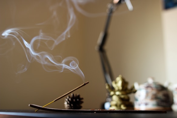 Wisps of smoke curl gracefully from a lit incense stick placed in a holder. The background features an unfocused area with a pine cone and a blurred decorative object, creating a serene and contemplative atmosphere.