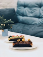 Two elegantly plated éclairs are placed on a white table, with one featuring a glossy chocolate glaze and caramel popcorn, and the other topped with colorful fruit and cream. A soft, plush couch in a muted blue tone serves as the background, lending a cozy atmosphere. A small potted plant sits nearby, adding a touch of greenery.
