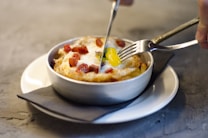 A fork and knife are slicing into a baked dish in a metal bowl. The dish features an egg with a runny yolk and appears to be topped with diced cooked bacon or sausage, possibly served over a bed of hash browns or similar ingredients. The bowl rests on a white plate and is placed over a dark napkin, creating a cozy dining experience.