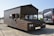 A unique vehicle designed to resemble a wooden shed with wheels, parked on an asphalt surface. The license plate reads 'Q710 LBW,' and the front features typical car parts such as headlights and a license plate. Nearby, there is a black van with 'MAD MAX RACE TEAM' written on it, along with some other vehicles partially visible in the background.
