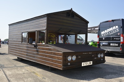 A unique vehicle designed to resemble a wooden shed with wheels, parked on an asphalt surface. The license plate reads 'Q710 LBW,' and the front features typical car parts such as headlights and a license plate. Nearby, there is a black van with 'MAD MAX RACE TEAM' written on it, along with some other vehicles partially visible in the background.