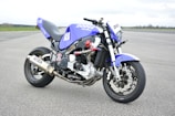 A custom motorcycle with a sporty and aggressive design displaying a blue and black color scheme. It features branding such as 'Dodge' and various stickers, with an exposed engine and metallic exhaust pipes. The bike is parked on a wide asphalt surface with a vast, open, and slightly cloudy sky in the background.
