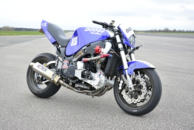 A custom motorcycle with a sporty and aggressive design displaying a blue and black color scheme. It features branding such as 'Dodge' and various stickers, with an exposed engine and metallic exhaust pipes. The bike is parked on a wide asphalt surface with a vast, open, and slightly cloudy sky in the background.