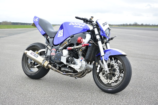 A custom motorcycle with a sporty and aggressive design displaying a blue and black color scheme. It features branding such as 'Dodge' and various stickers, with an exposed engine and metallic exhaust pipes. The bike is parked on a wide asphalt surface with a vast, open, and slightly cloudy sky in the background.