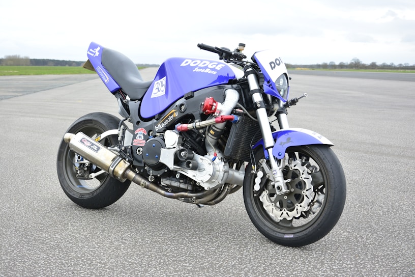A custom motorcycle with a sporty and aggressive design displaying a blue and black color scheme. It features branding such as 'Dodge' and various stickers, with an exposed engine and metallic exhaust pipes. The bike is parked on a wide asphalt surface with a vast, open, and slightly cloudy sky in the background.