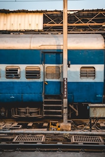 A blue and white train car is parked at a station, with windows featuring protective grills. A metal pole stands prominently in front of the train, partially obscuring a door with a small staircase leading up to it. Overhead, a metal roof structure is visible, with pipes and wiring scattered below, indicating a functional and industrial environment.