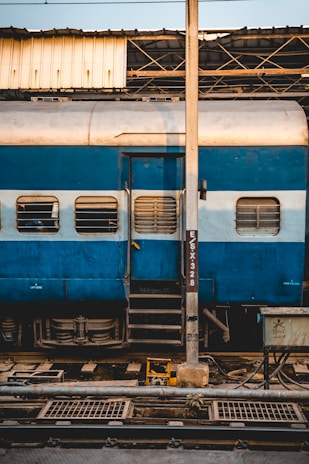 A blue and white train car is parked at a station, with windows featuring protective grills. A metal pole stands prominently in front of the train, partially obscuring a door with a small staircase leading up to it. Overhead, a metal roof structure is visible, with pipes and wiring scattered below, indicating a functional and industrial environment.
