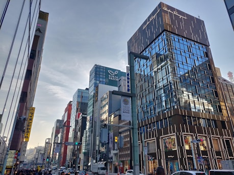 A bustling city street featuring modern, tall buildings with reflective glass facades under a partly cloudy sky. Various brand logos are visible on the buildings, and people and vehicles are on the street. The busy urban environment suggests commercial activity and movement.