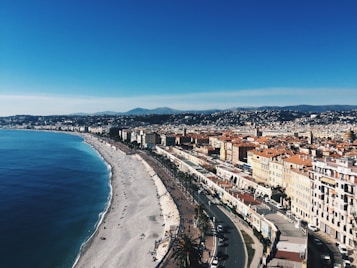 A coastal cityscape with a sweeping view of a curved beach bordered by turquoise water on one side and a row of buildings with red-tiled roofs on the other. A promenade runs parallel to the beach, lined with palm trees and people enjoying the sunny day. In the background, gently rolling hills are visible under a clear blue sky.