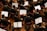 Musicians in formal black attire are seated in rows, playing violins while looking at sheet music in front of them. The scene is a concert or orchestra setting with wooden floors and red chairs.