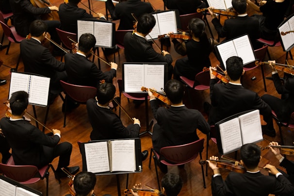Musicians in formal black attire are seated in rows, playing violins while looking at sheet music in front of them. The scene is a concert or orchestra setting with wooden floors and red chairs.