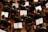 Musicians in formal black attire are seated in rows, playing violins while looking at sheet music in front of them. The scene is a concert or orchestra setting with wooden floors and red chairs.