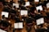 Musicians in formal black attire are seated in rows, playing violins while looking at sheet music in front of them. The scene is a concert or orchestra setting with wooden floors and red chairs.